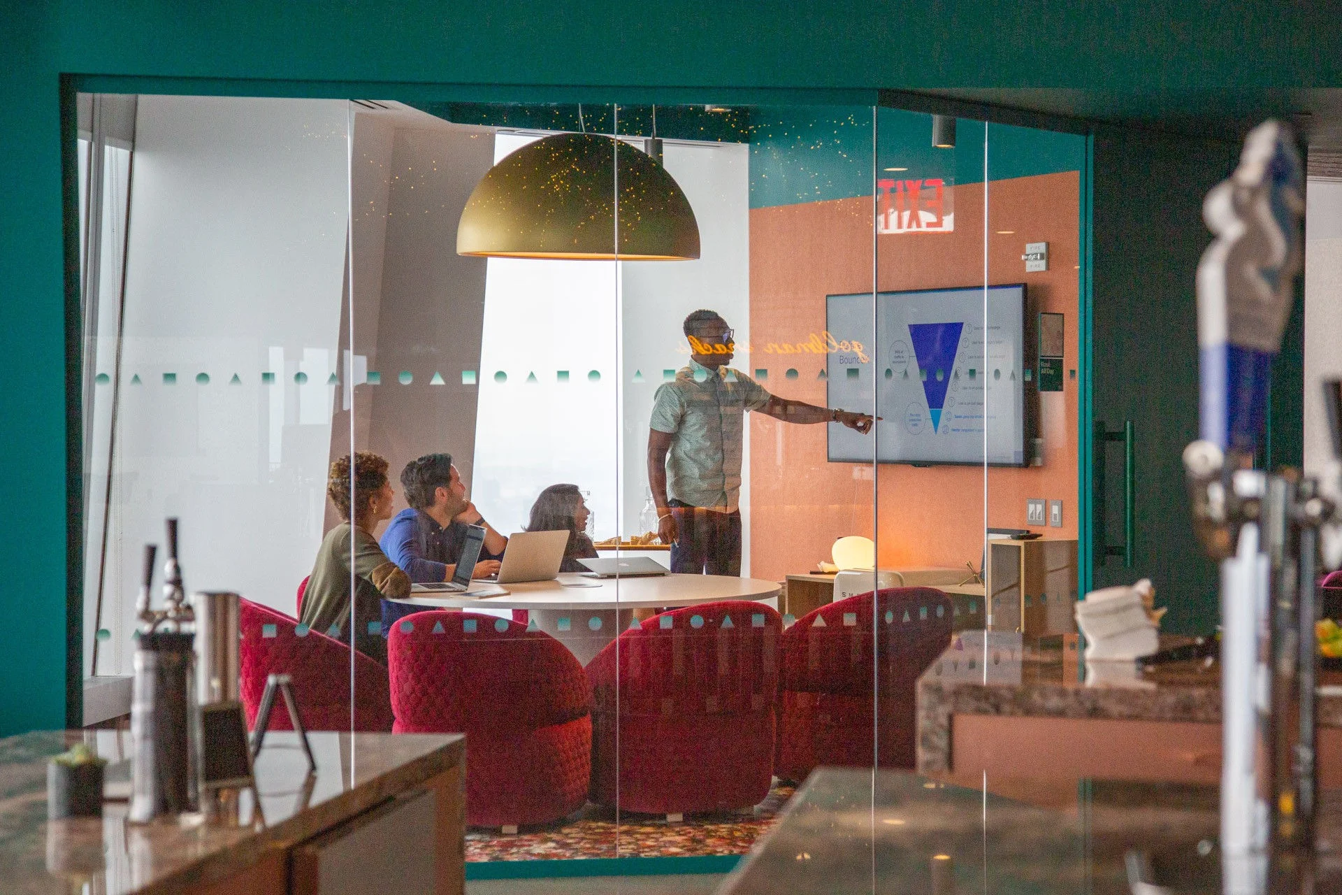 Team meeting in a modern office, with a presenter pointing at a screen while colleagues sit around a table.
