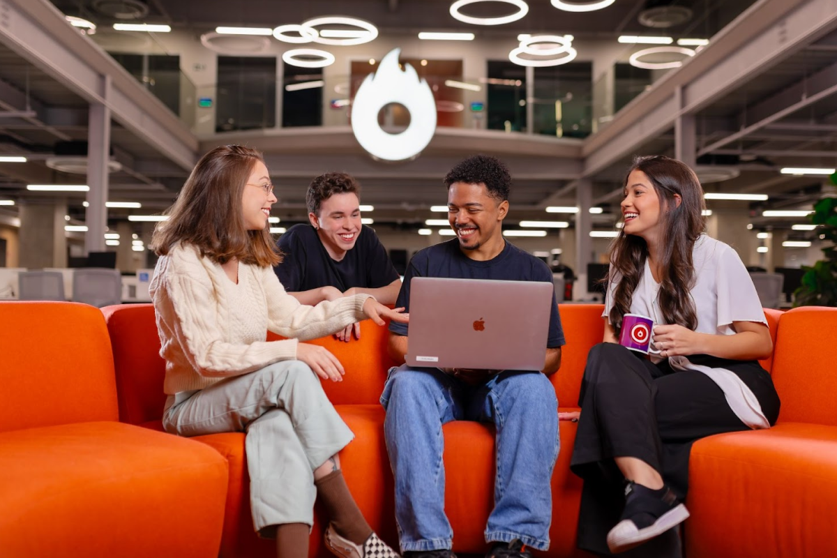 A diverse group of four smiling employees sitting on a bright orange sofa in a modern office, gathered around a laptop. A large, illuminated Hotmart logo is visible in the background.