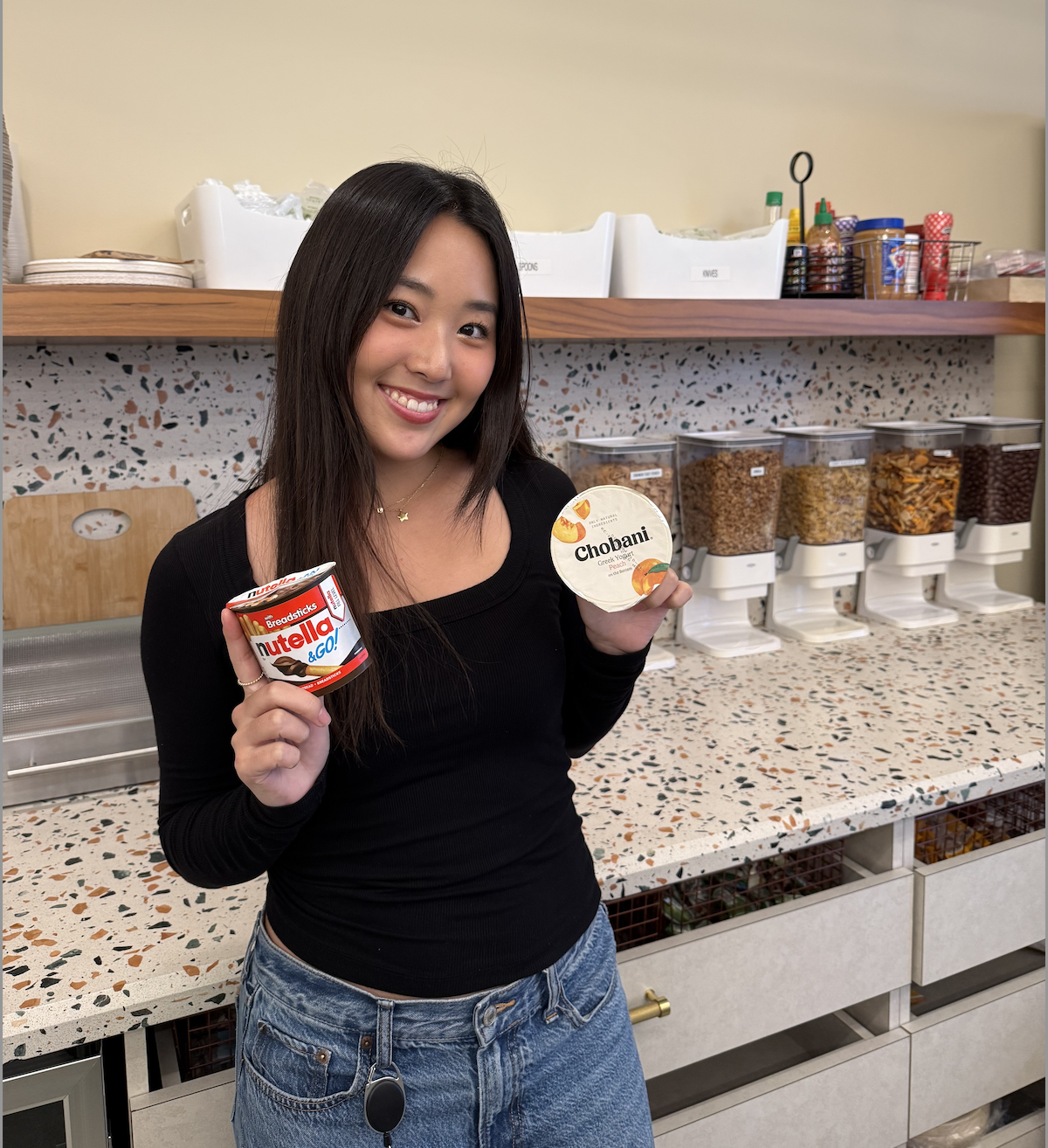 A woman in a black top and jeans is holding a Nutella cup in one hand and a Chobani yogurt cup in the other. 